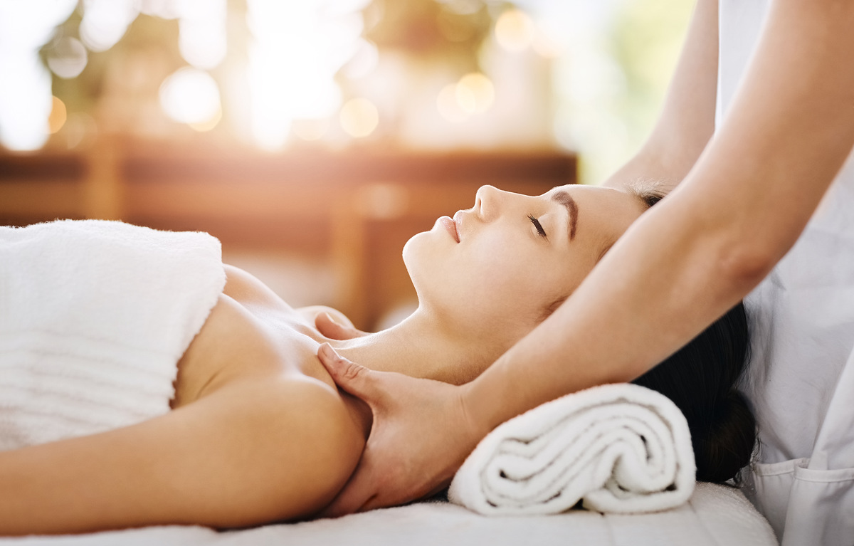A woman wearing glasses is lying down with a comfortable expression, as the massage therapist's hands gently handle the face and neck area