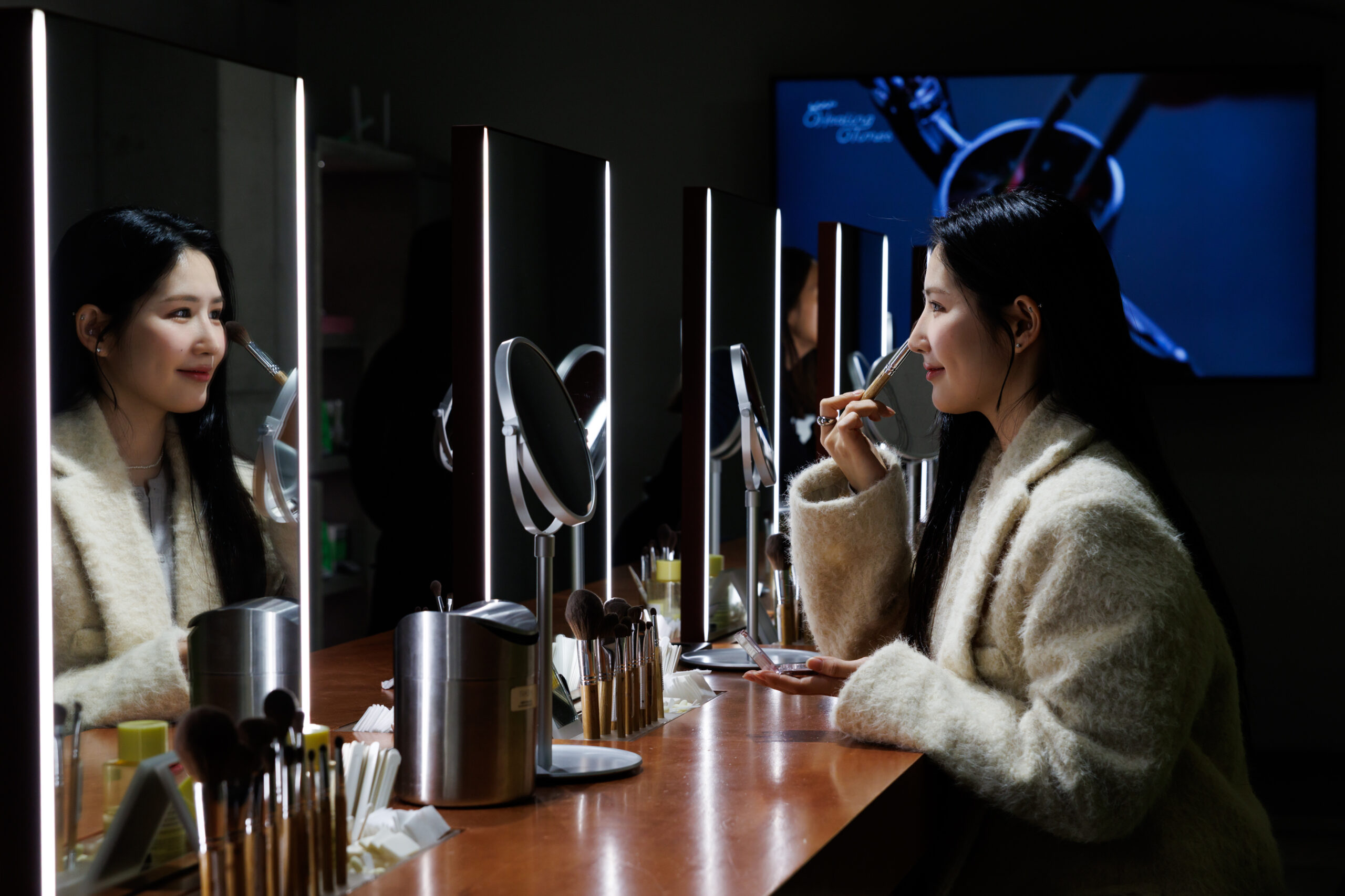 A woman's back view as she follows her beauty routine in front of a lighted mirror