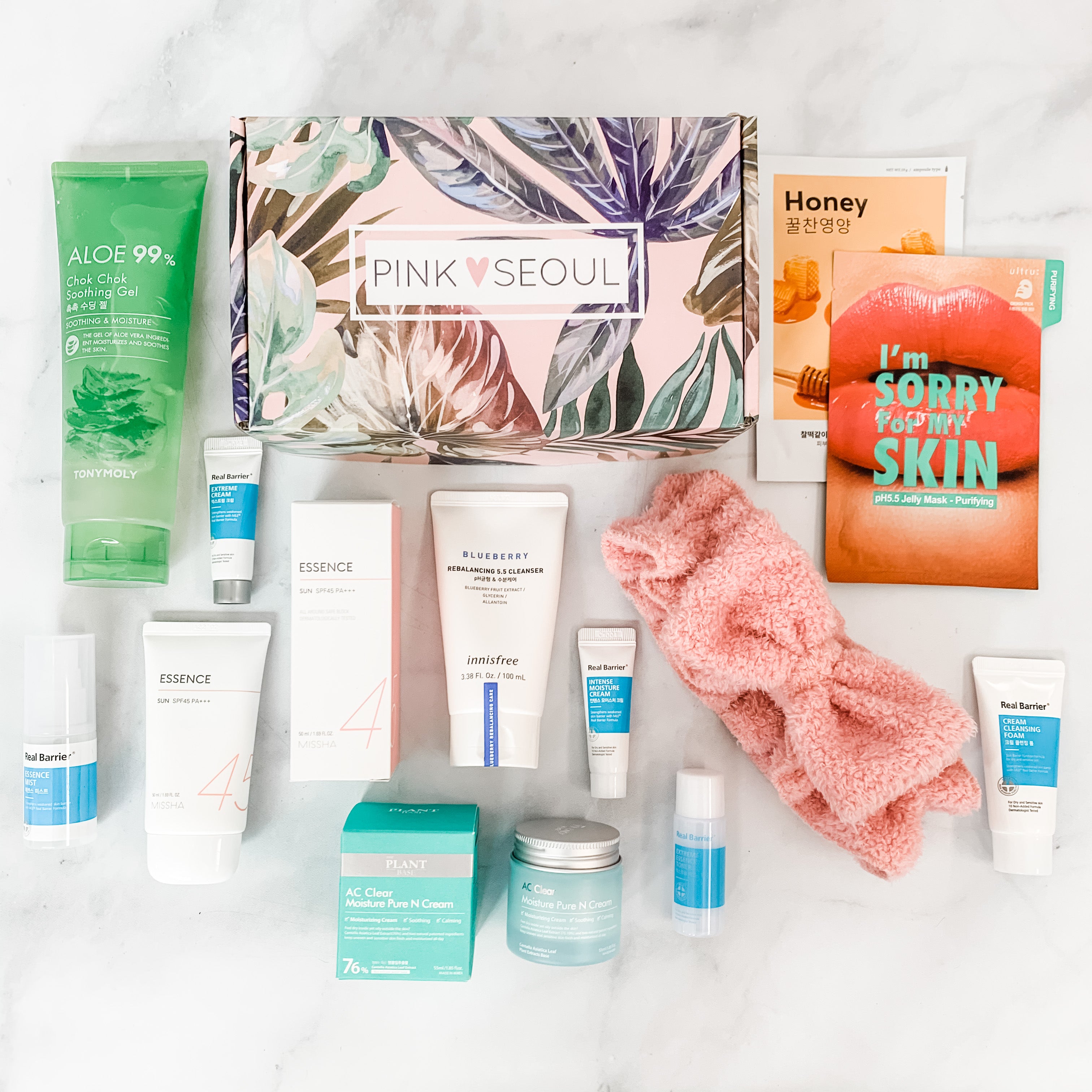 A scene with skincare products such as aloe gel, cleanser, essence, facial mask sheets, and moisturizer, placed in front of a pink leaf-pattern box, along with a pink towel