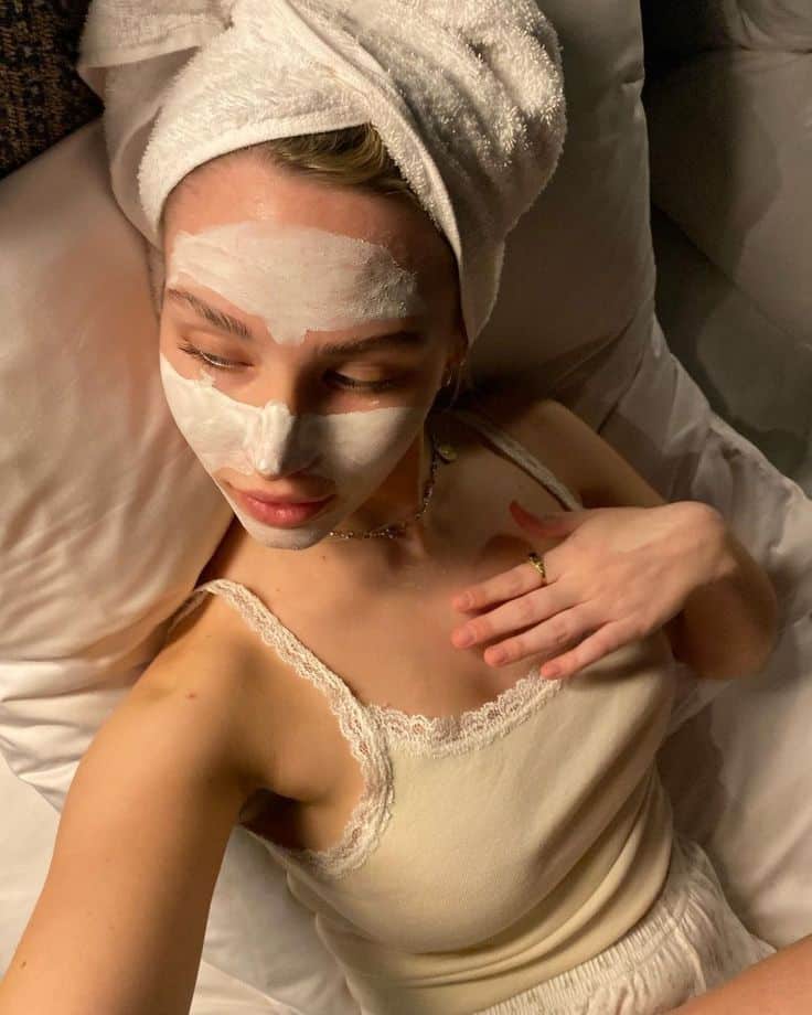 A woman wrapped in a white towel and applying a white sleep mask resting on a bed